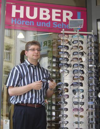 Mann vor Optikergeschäft mit Sonnenbrillenständer, Schild „Hören und Sehen HUBER“.
